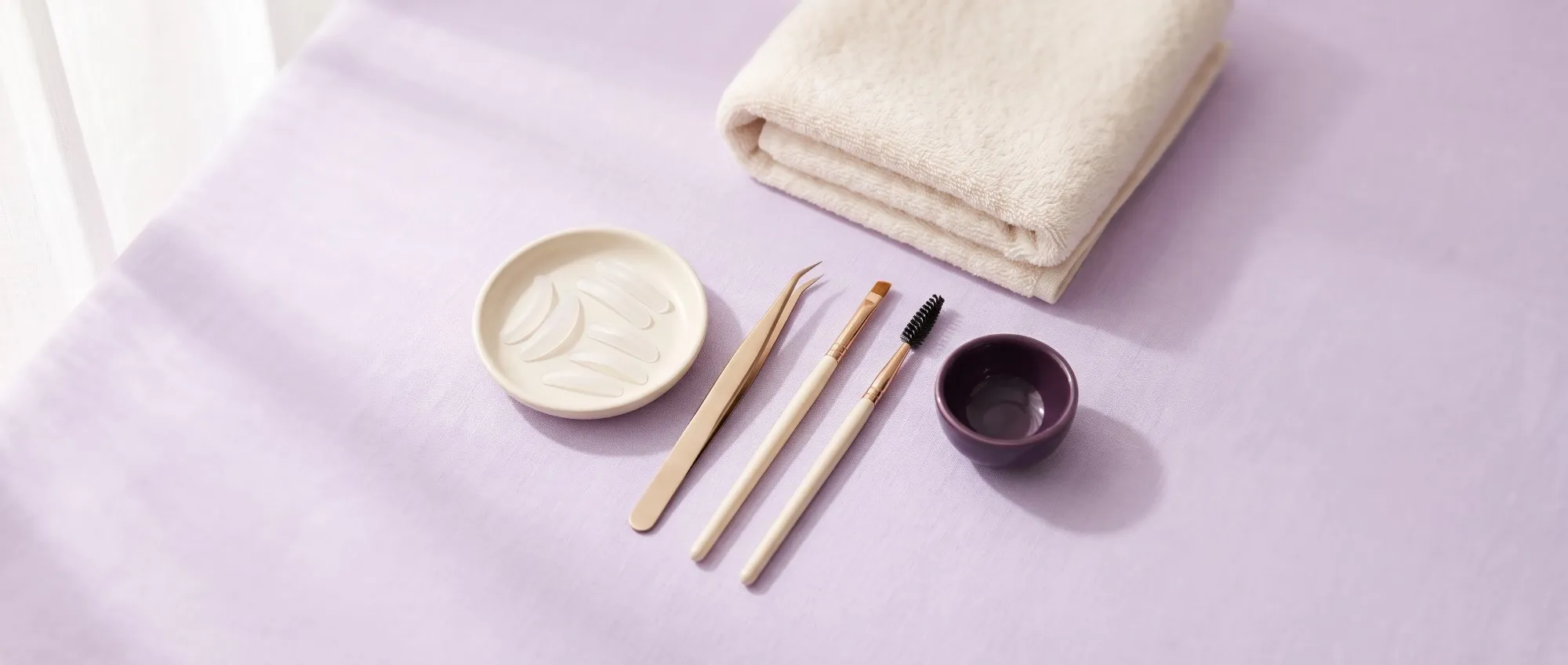 Kit de lifting de pestañas: almohadillas de silicona en un platito, pinza, pincel fino y cepillito, junto a una toalla crema sobre fondo lavanda