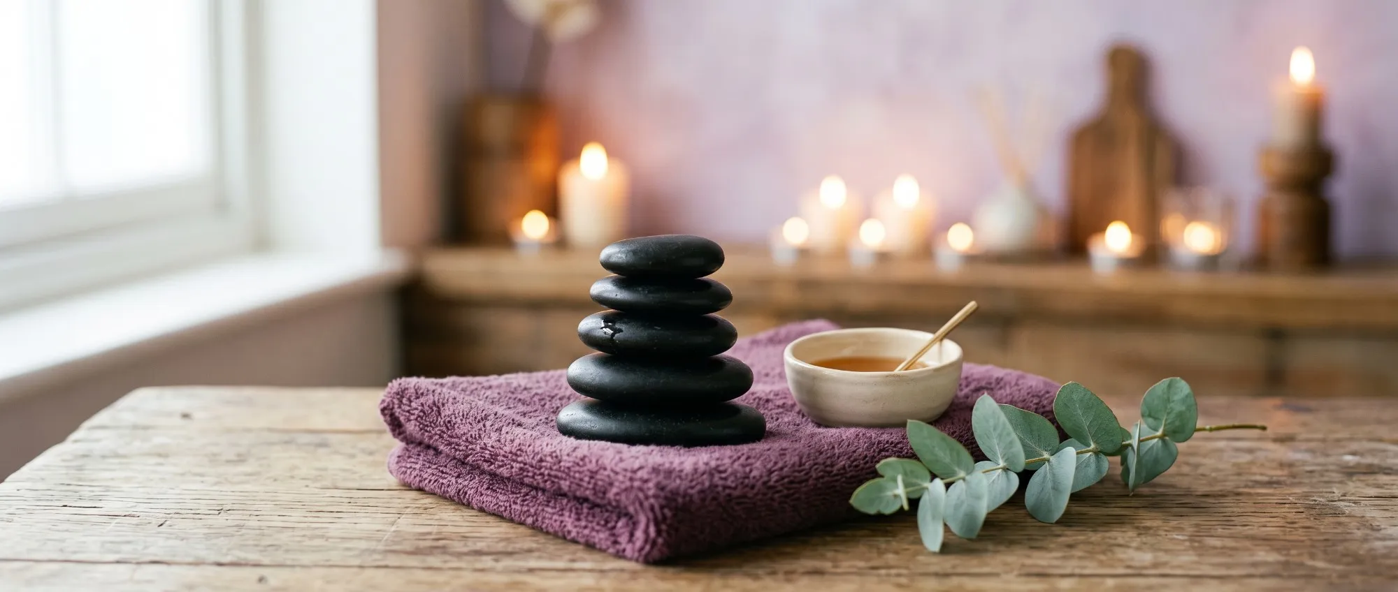 Piedras calientes apiladas sobre una toalla color ciruela, con un bowl de aceite para masajes, ramas de eucalipto y velas encendidas de fondo